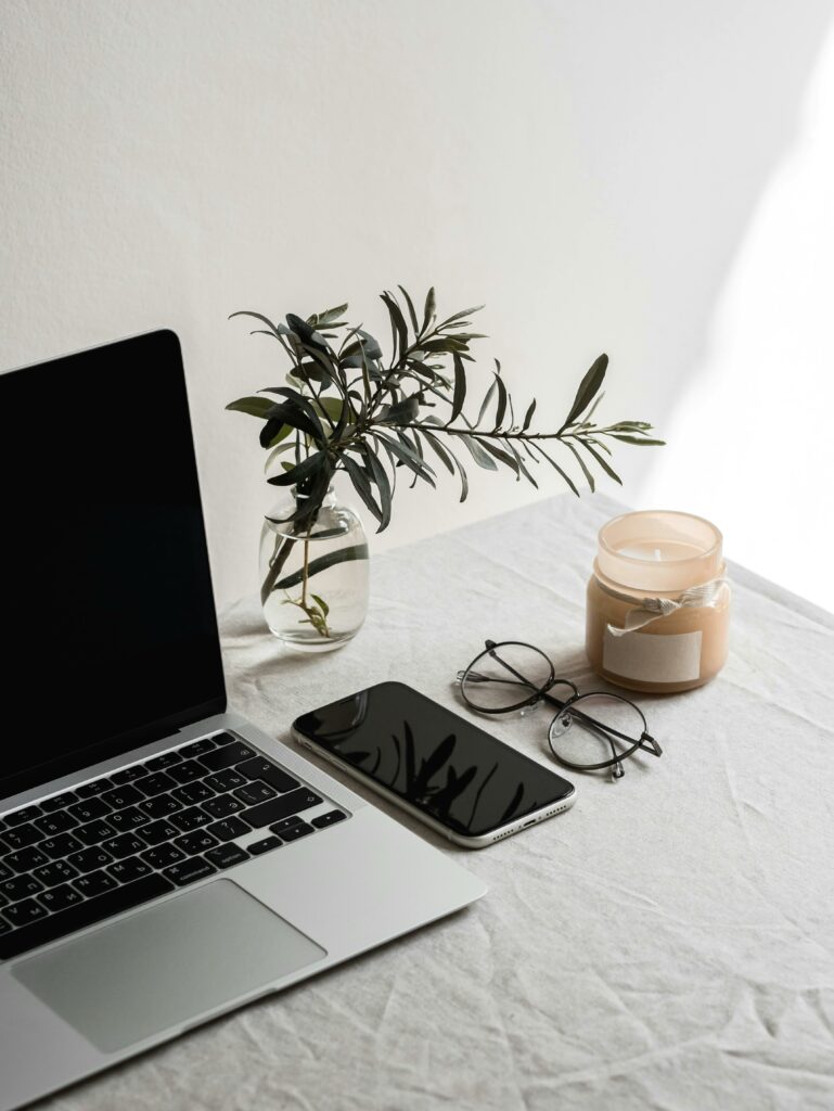A minimalist indoor workspace setup with a laptop, smartphone, eyeglasses, candle, and plant.